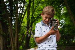 Kindergartenfotograf Weissenhorn. Ein Junge schaut angestrengt durch eine sehr alte Kamera. Im Hintergrund schön grün