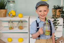 Ein Junge mit Boxermütze und Hosenträgern steht vor einem modernen Kindergartenfoto-Set. Er hält einen Limonadenglas in der Hand